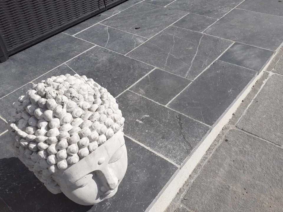 Scène extérieure de chantier urbain où un ouvrier en vêtements noirs et gants jaunes de protection pose des pavés autobloquants gris sur une surface gravillonnée